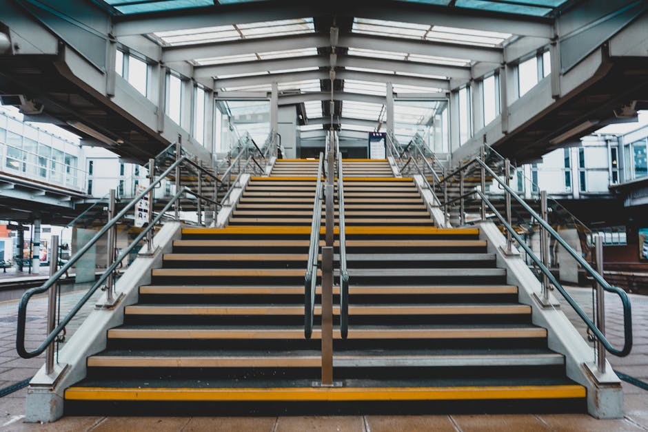 A set of wide outdoor stairs with black rubber anti-slip strips and yellow safety edges, leading up to the entrance of East Croydon station. The stairs are flanked by metal handrails on both sides, with additional handrails in the middle dividing the staircase into two sections. The station features a modern glass and steel structure with a transparent roof allowing natural light to illuminate the area. At the top of the stairs, there is a covered walkway with glass panels and metal framing, providing access to the station platform. In the background, there are glimpses of surrounding urban buildings and a bus stop to the left. The scene captures a clean, organized, and well-lit environment suitable for pedestrian movement and station access, relevant to house removals and furniture transport logistics for relocation services like those offered by Man with Van Croydon.
