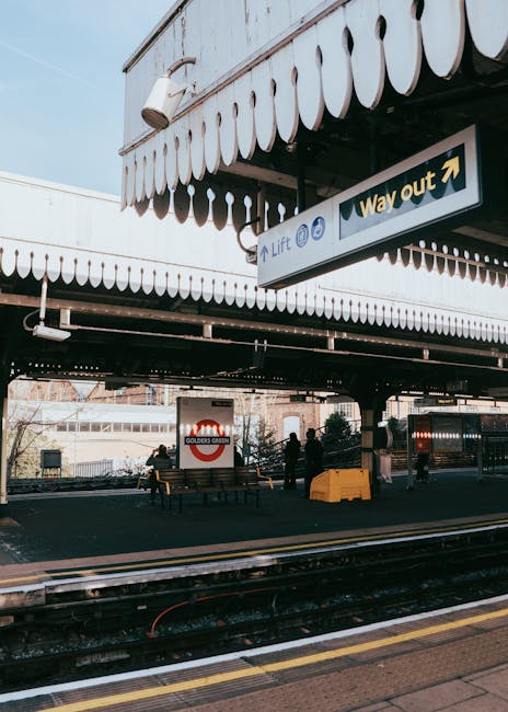A view of East Croydon station platform with a canopy featuring decorative scalloped trim and a large sign indicating the way out. Under the canopy, there are several people standing and sitting on benches, with some engaging in activities related to home relocation or packing logistics. The platform shows multiple dark-colored benches and a yellow plastic container, with a backdrop of train tracks and nearby station structures. The scene suggests preparations for furniture transport or moving services, with environmental lighting indicating daytime. The station environment, combined with the presence of movable items and people, reflects the context of house removals and moving operations facilitated by Man with Van Croydon.