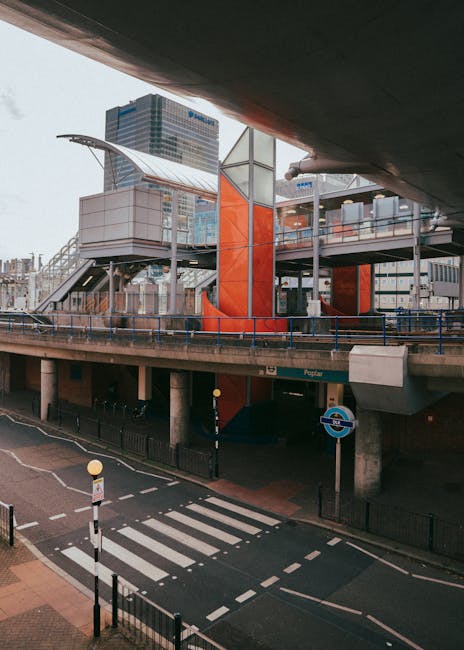 A set of wide outdoor stairs with black rubber anti-slip strips and yellow safety edges, leading up to the entrance of East Croydon station. The stairs are flanked by metal handrails on both sides, with additional handrails in the middle dividing the staircase into two sections. The station features a modern glass and steel structure with a transparent roof allowing natural light to illuminate the area. At the top of the stairs, there is a covered walkway with glass panels and metal framing, providing access to the station platform. In the background, there are glimpses of surrounding urban buildings and a bus stop to the left. The scene captures a clean, organized, and well-lit environment suitable for pedestrian movement and station access, relevant to house removals and furniture transport logistics for relocation services like those offered by Man with Van Croydon.