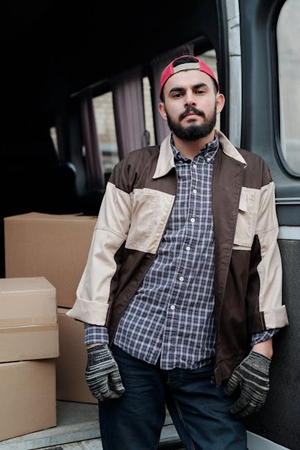 A male mover with a beard, dressed in a white polo shirt and black cap, stands outside a moving van holding a cardboard box during a home relocation. The van's rear doors are open, revealing multiple 